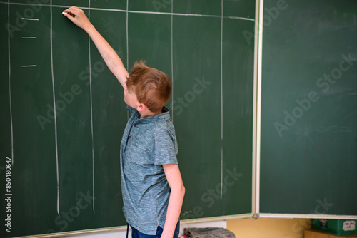 Young student is writing on a large green chalkboard while solving a math problem