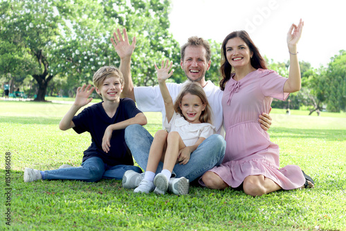 Happy family sitting on green grass and waving hands at park, smiling father, mother, son and daughter spending time together outdoors in beautiful garden on holiday. Parents and kids have fun outside