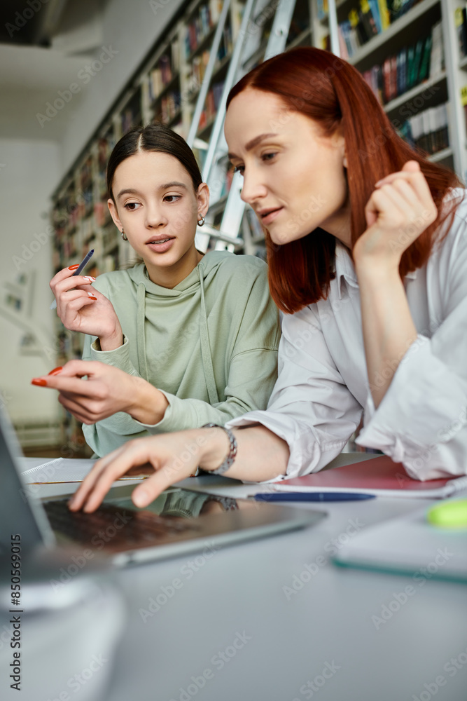 A tutor with red hair guides a teenage student in a library, both engrossed in the laptop screen for after-school lessons. A tutor with red hair guides a teenage student in a library, both engrossed in the laptop screen for after-school lessons.
