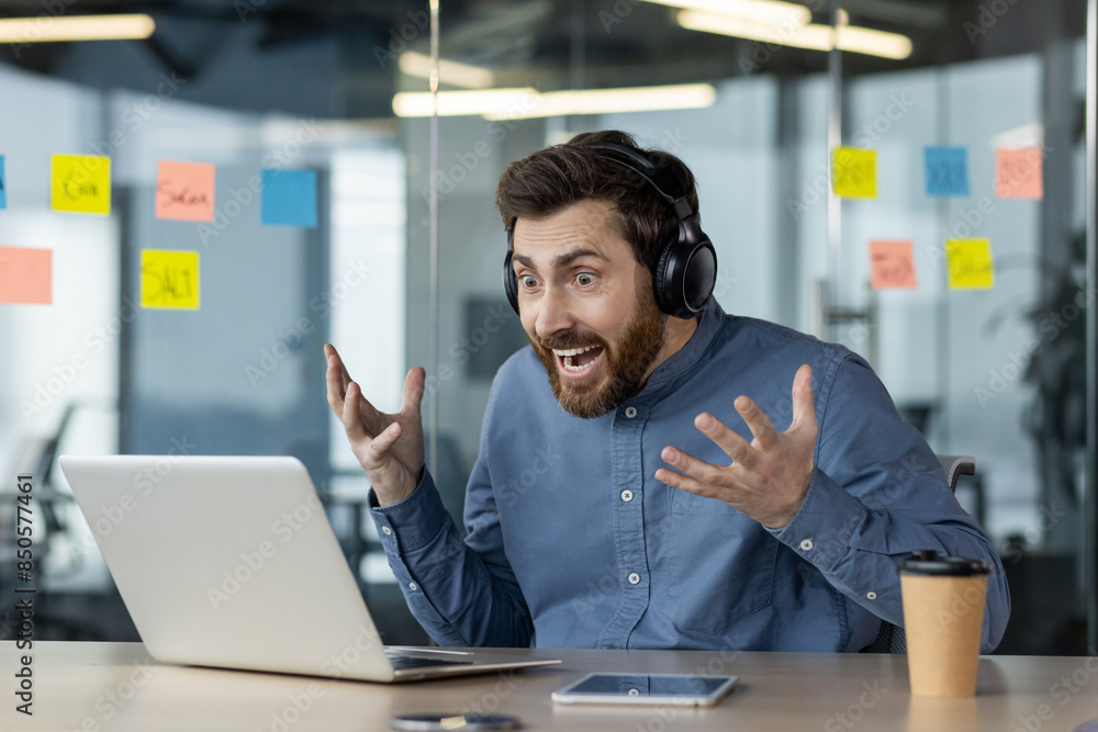 Surprised young man sitting at desk in office wearing headphones and looking shocked at laptop screen Surprised young man sitting at desk in office wearing headphones and looking shocked at laptop screen
