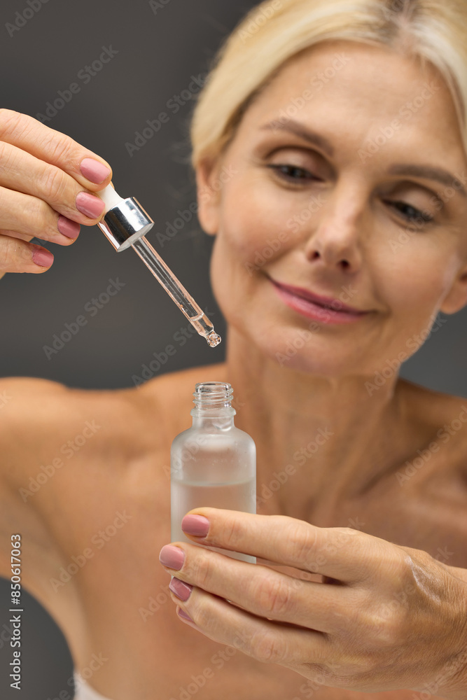 Charming mature woman applying serum on a gray backdrop. Charming mature woman applying serum on a gray backdrop.