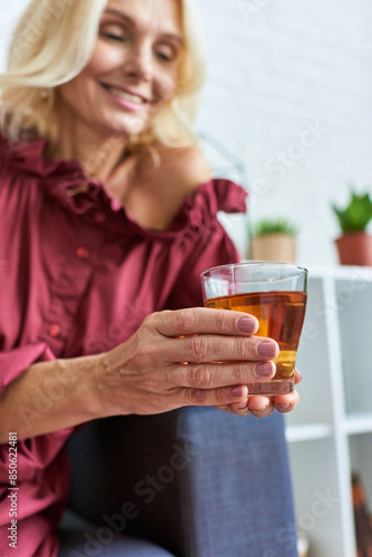 Mature woman in stylish dress relaxes on couch, holding glass of tea.