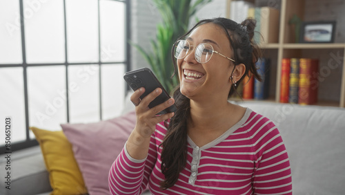 Laughing young hispanic woman enjoying a conversation on her smartphone at home, surrounded by a cozy interior. photo