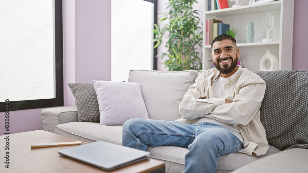 A cheerful bearded man sits crossed-arms on a couch in a modern living room, radiating confidence and comfort. A cheerful bearded man sits crossed-arms on a couch in a modern living room, radiating confidence and comfort.