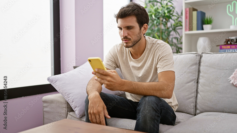 Handsome hispanic man using smartphone while sitting on couch at home, exuding a sense of casual lifestyle and connectivity. Handsome hispanic man using smartphone while sitting on couch at home, exuding a sense of casual lifestyle and connectivity.