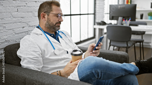 Quadro su tela Handsome mature man with grey hair in doctor's coat holding coffee and smartphone indoors at clinic workplace