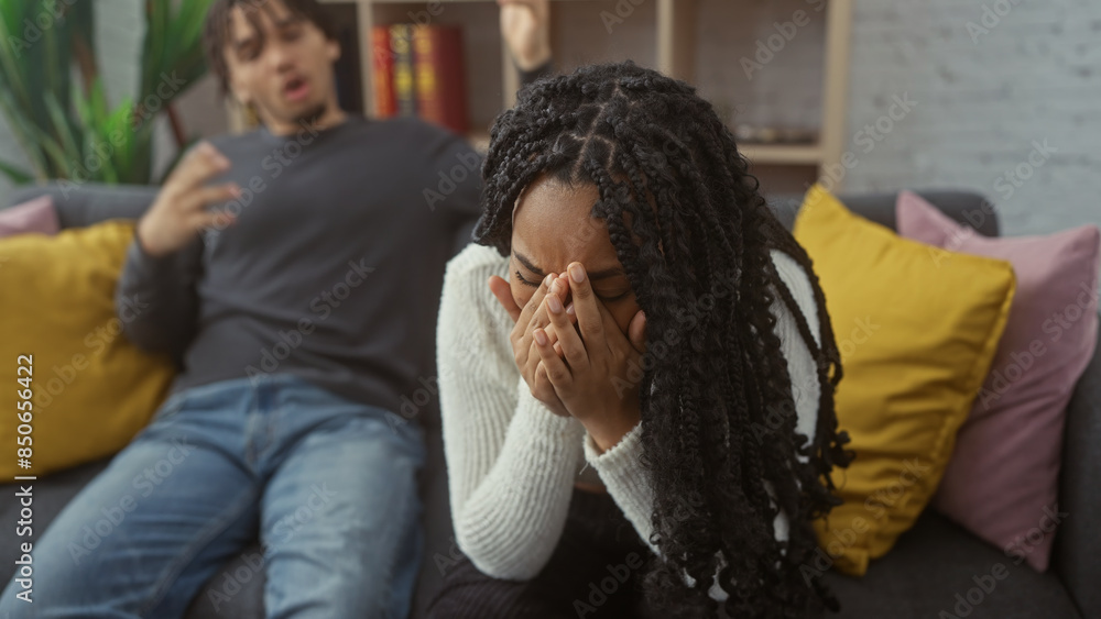 Fototapeta premium A distressed woman covers her face with hands beside a frustrated man on a couch, depicting a couple's argument in a living room.