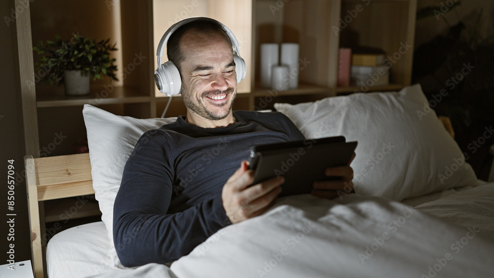 Smiling bearded man using tablet with headphones in bed at night, reflecting modern leisure or relaxation at home. Smiling bearded man using tablet with headphones in bed at night, reflecting modern leisure or relaxation at home.