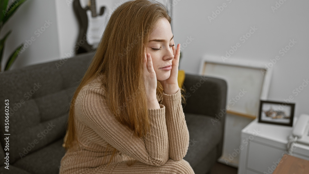 A serene young woman sitting with eyes closed and hands on her cheeks in a cozy living room, evoking a sense of calm.