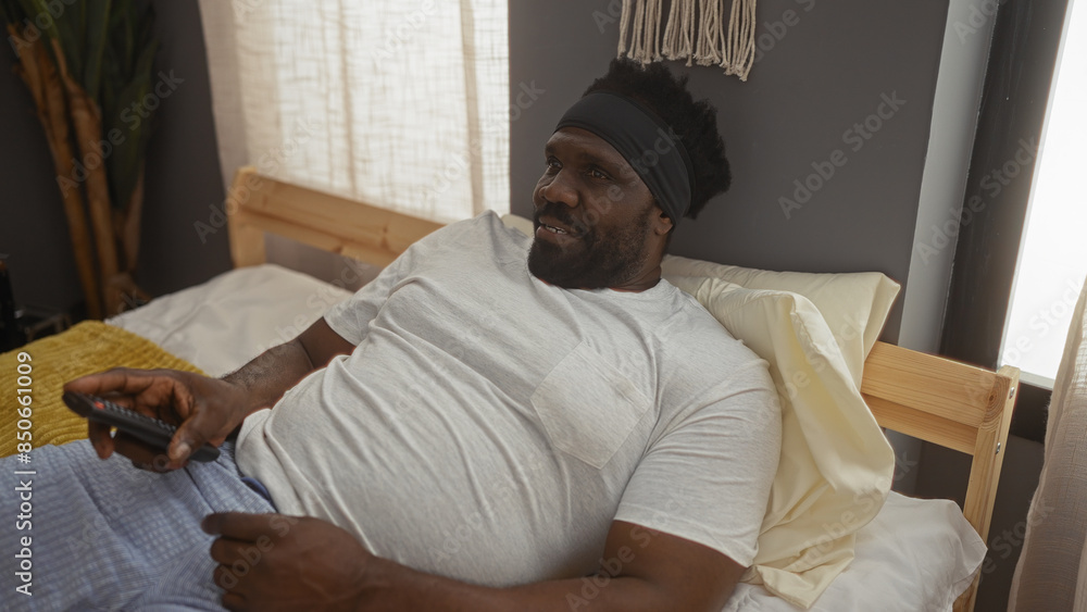 Fototapeta premium A young, handsome african american man with a beard relaxes in bed at home, holding a remote, in a cozy and well-lit bedroom.