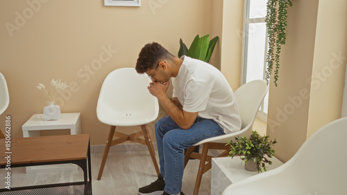 A young, hispanic man is seated in a waiting room with a thoughtful expression, surrounded by white chairs and indoor plants. © Krakenimages.com