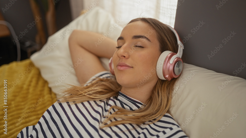 A young attractive blonde woman lounges in bed wearing headphones, enjoying music in the cozy indoor setting of her bedroom at home. A young attractive blonde woman lounges in bed wearing headphones, enjoying music in the cozy indoor setting of her bedroom at home.
