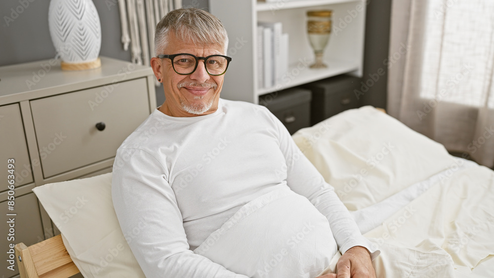 Smiling middle-aged man with glasses reclining comfortably in a bright bedroom setting, exuding a sense of relaxation and contentment. Smiling middle-aged man with glasses reclining comfortably in a bright bedroom setting, exuding a sense of relaxation and contentment.