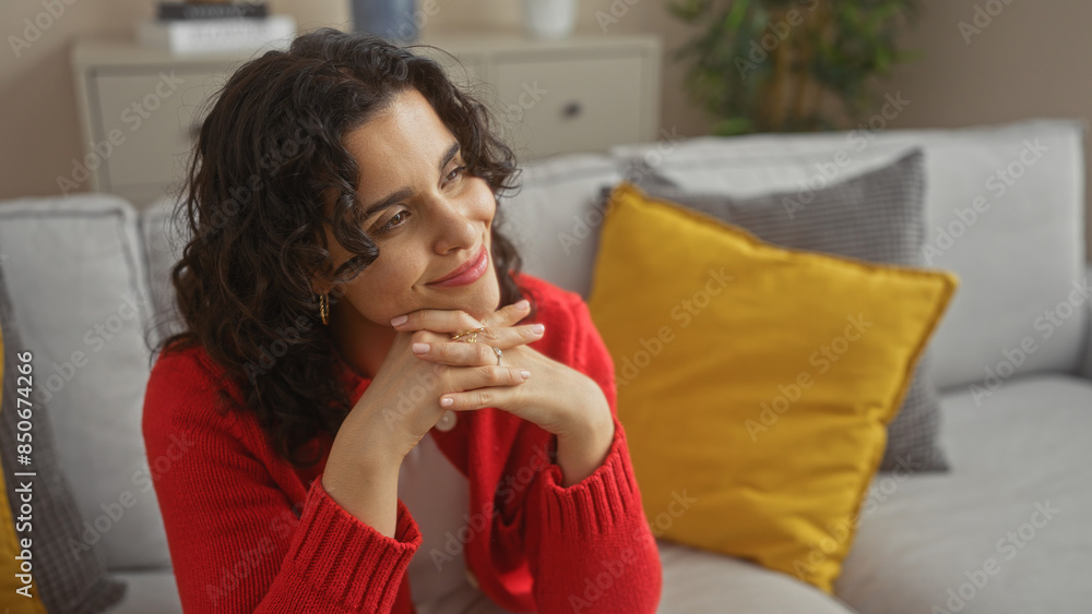 Beautiful adult hispanic brunette woman sitting in a cozy apartment living room wearing a red sweater, with her hands resting beneath her chin, thoughtful and smiling. Beautiful adult hispanic brunette woman sitting in a cozy apartment living room wearing a red sweater, with her hands resting beneath her chin, thoughtful and smiling.