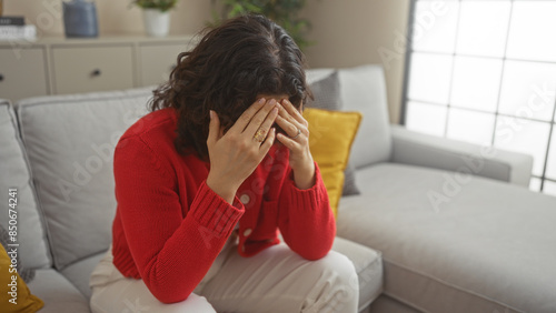 Obraz na plátně A young hispanic woman in a red sweater sits in a modern living room, covering her face with her hands in apparent distress