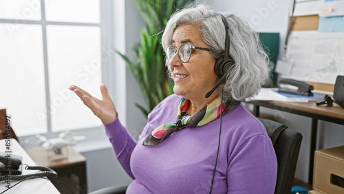 Cheerful mature woman with grey hair wearing glasses and headset talking in a modern office. photo