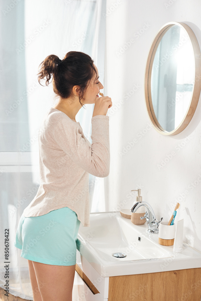 A brunette woman brushes her teeth in front of a bathroom mirror in morning time A brunette woman brushes her teeth in front of a bathroom mirror in morning time