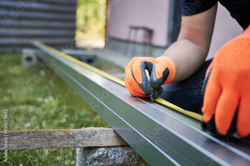 Close up of man hands in work gloves using tape measure and drawing mark on metal rail with marker pen. Male worker measuring and marking aluminum mounting rail for solar panel installation.