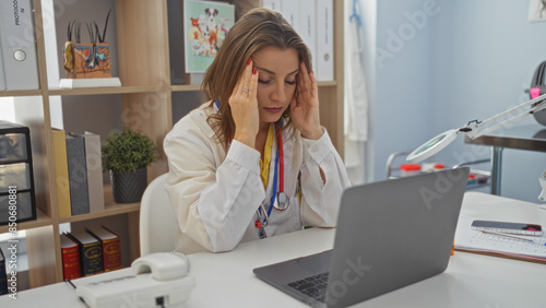 A young attractive blonde woman working in a vet clinic appears stressed while seated at a desk with a laptop in an indoor office environment, surrounded by medical equipment and documents.