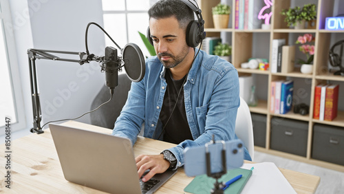 Handsome hispanic man podcasts from a modern studio wearing headphones and using laptop and microphone