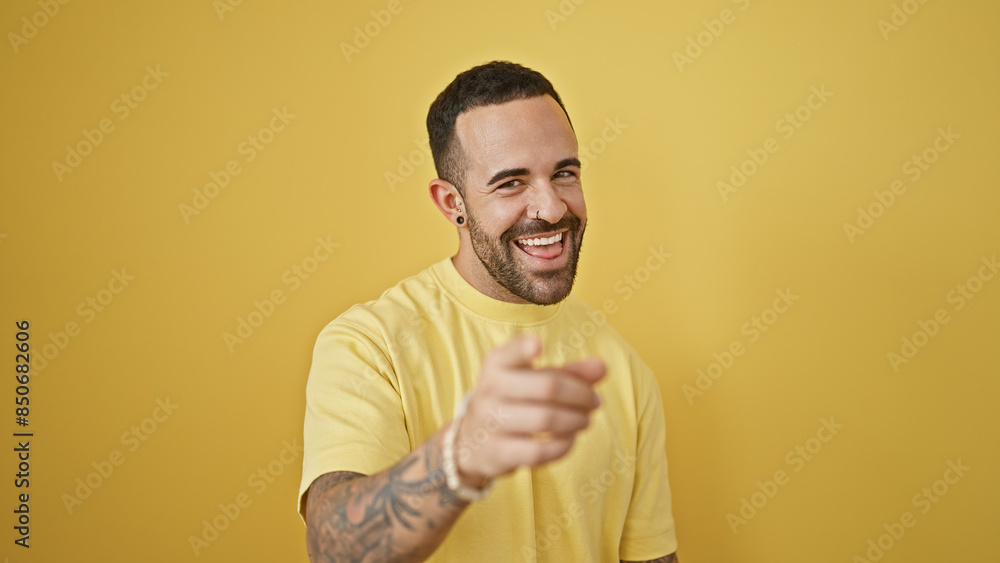 A cheerful young hispanic man with a beard, smiling and pointing, isolated against a yellow wall background. A cheerful young hispanic man with a beard, smiling and pointing, isolated against a yellow wall background.