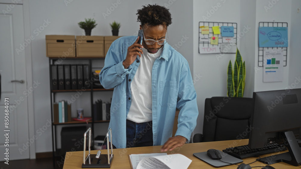 Fototapeta premium Young african american man with a beard talking on the phone while working at his desk in a modern office setting.