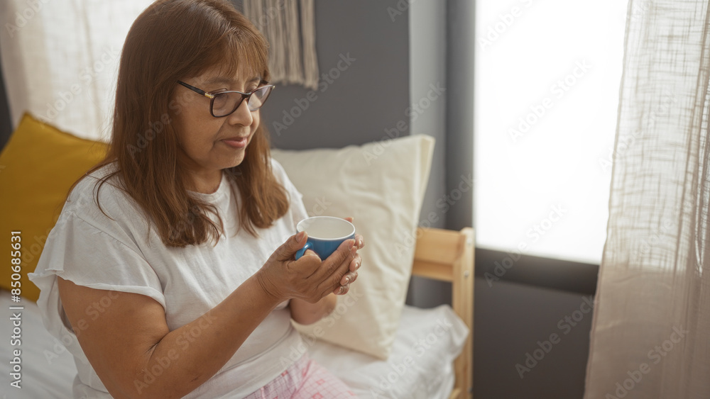 A mature hispanic woman sits indoors in her bedroom, holding a cup while wearing glasses and pajamas on a cozy bed. A mature hispanic woman sits indoors in her bedroom, holding a cup while wearing glasses and pajamas on a cozy bed.