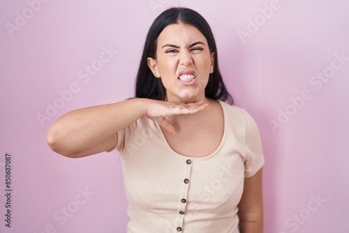 Young hispanic woman standing over pink background cutting throat with hand as knife, threaten aggression with furious violence