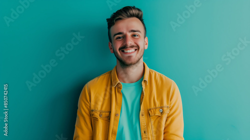A confident millennial man in a casual shirt, smiling brightly at the camera against a solid teal background