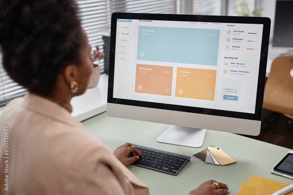 Over shoulder shot of African American woman using computer in office. PC screen displaying online education platform with UI, UX design lessons, focus on monitor, copy space Over shoulder shot of African American woman using computer in office. PC screen displaying online education platform with UI, UX design lessons, focus on monitor, copy space