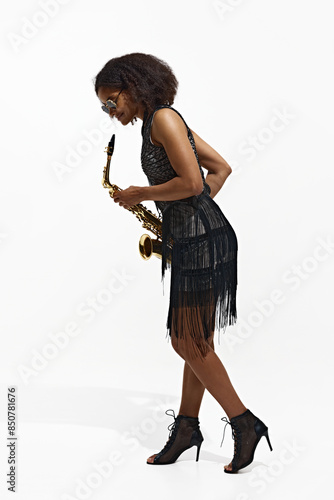Photography Full-length portrait of African-American young woman in black dress and sunglasses standing with saxophone isolated on white background