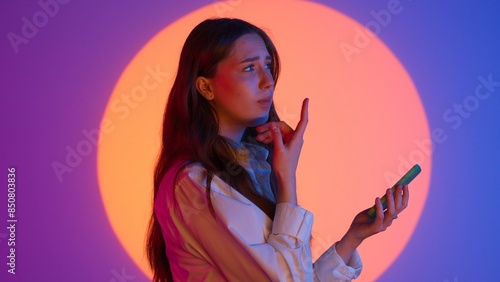 Stylish young woman in casual clothing holds smartphone, hesitates sending text message, confused expression. Isolated on colorful studio background. photo