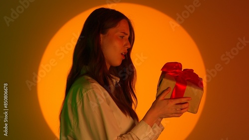 Stylish young woman in casual clothing holding gift box, shaking the box thinks what's inside. Isolated on colorful studio background yellow spotlight. photo