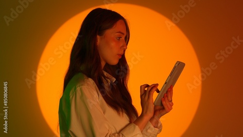 Stylish young woman in casual clothing using tablet, scrolling swiping online social media. Isolated on colorful studio background yellow spotlight. photo