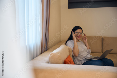 Foto Pleased Asian lady with laptop talking on mobile phone