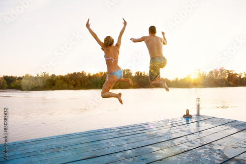 Young friends jumping into the water from jetty. Rear view of young couple having fun at the lake on summer day. Summer holidays.