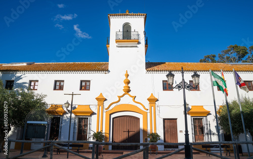 a traditional Spanish building with a white facade and distinctive yellow architectural details, featuring a bell tower, under a clear blue sky, flanked by the Spanish and Andalusian flags photo