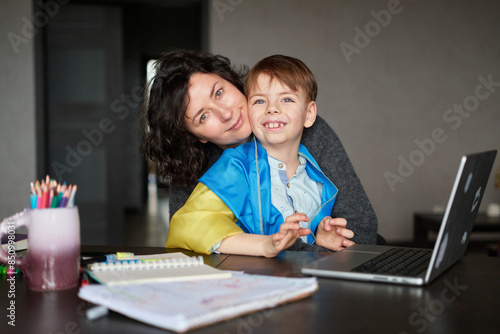 Ukrainian mother and son with flag sitting with laptop at home studying .Happy smiling family hugs and looking at the camera. Ukrainian refugees concept. High quality photo