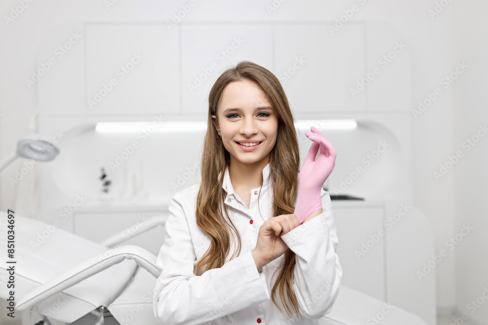 Young Woman Nurse Putting on a Pink Rubber Glove. White Doctor's Coat. White Hospital Office. Smile. Young Woman Nurse Putting on a Pink Rubber Glove. White Doctor's Coat. White Hospital Office. Smile.