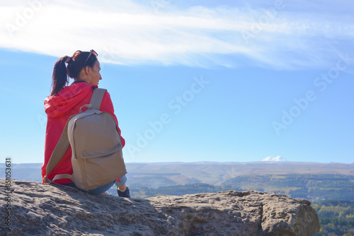 Obraz na płótnie A girl tourist with a backpack sits on the top of a mountain and enjoys the beautiful views. A walking woman in an orange jacket rests on a rock, looking at the beautiful sunny landscape of the w salonie