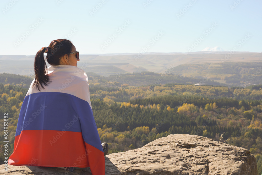 A girl tourist with a Russian flag on her shoulders sits on the top of a mountain and enjoys the beautiful views of the Caucasus Mountains.