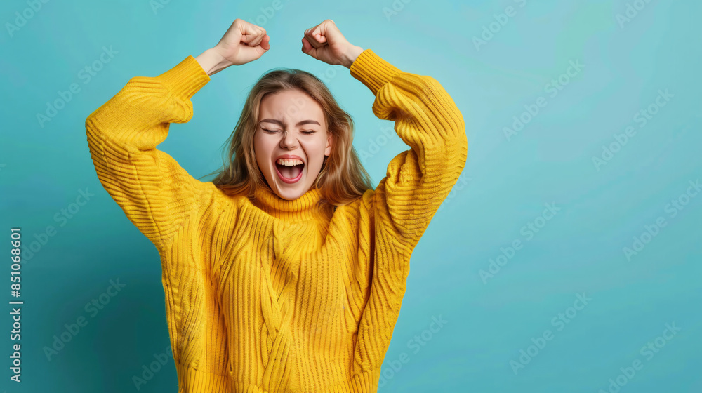 Excited young woman in a yellow sweater cheering with raised fists against a blue background Excited young woman in a yellow sweater cheering with raised fists against a blue background