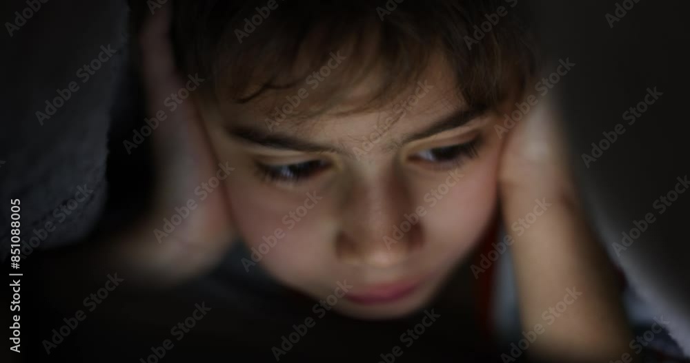 A close-up shot of a boy engrossed in his digital tablet, illuminated by its soft glow in the darkness of his room. A close-up shot of a boy engrossed in his digital tablet, illuminated by its soft glow in the darkness of his room.