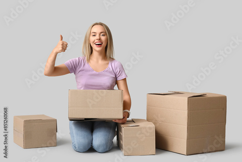 Happy young woman with cardboard boxes showing thumb-up on grey background. Moving into new house concept