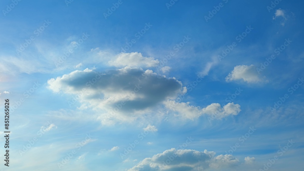 Obraz premium A serene blue sky with a scattering of fluffy white clouds. The sunlight gently illuminates the clouds, casting subtle shadows and creating a peaceful, picturesque scene. Blue sky background. 