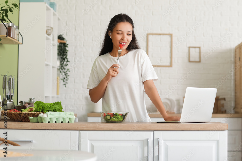 Beautiful young happy Asian woman with laptop and bowl of fresh vegetable salad in kitchen Beautiful young happy Asian woman with laptop and bowl of fresh vegetable salad in kitchen