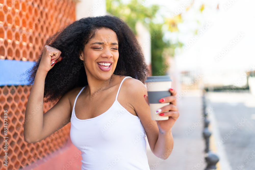 Young African American woman holding a take away coffee at outdoors celebrating a victory Young African American woman holding a take away coffee at outdoors celebrating a victory
