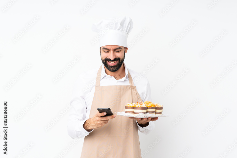 Young man holding muffin cake over isolated white background sending a message with the mobile Young man holding muffin cake over isolated white background sending a message with the mobile