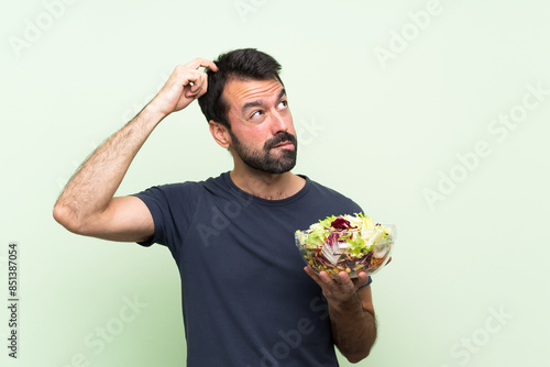 Young handsome man with salad over isolated green wall having doubts while scratching head photo