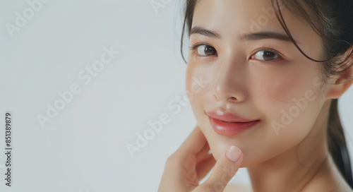 In a skincare commercial studio shoot, a beautiful Asian woman smiles gracefully with her hand resting on her cheek against a pristine white background, illuminated by studio lighting.